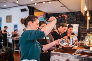 People are making their own chocolate bars at Wellington Chocolate Factory. A person is using a ladle to mix melted chocolate in a silver bowl.