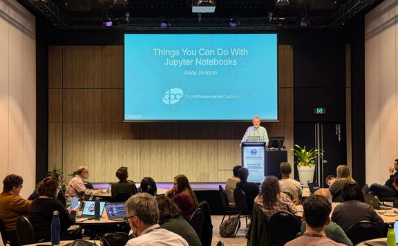 An audience sits in a conference room watching a speaker at a podium presenting a slide titled “Things You Can Do With Jupyter Notebooks.”