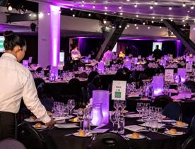 Inside the member's gallery of the Sky Stadium Function centre, many tables around the room all lit up with purple LED centre pieces, water, glasses, and plates with snacks. A waiter stands in the foreground serving plates.
