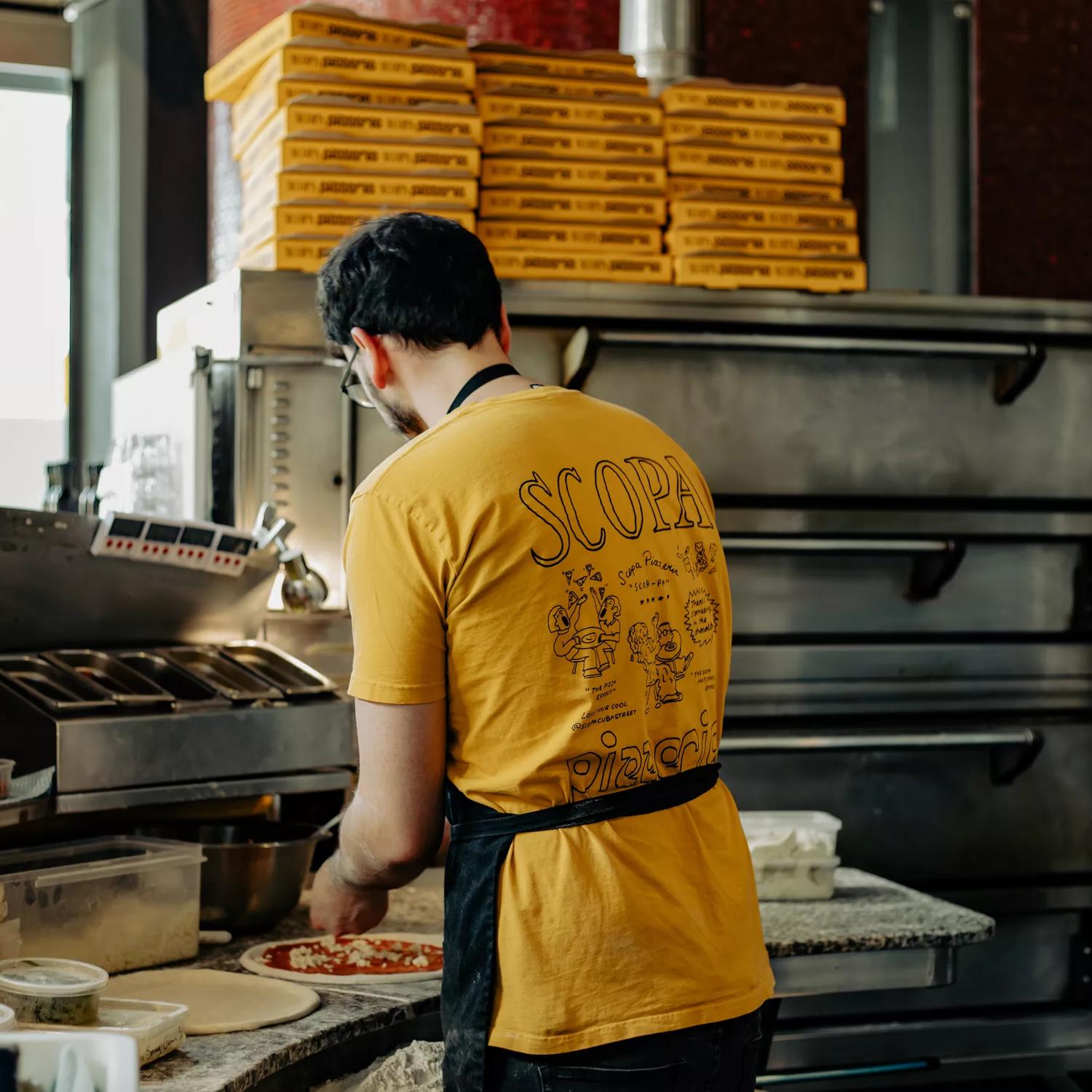 A chef from Scopa making pizzas with their back turned, wearing a yellow Scopa shirt.
