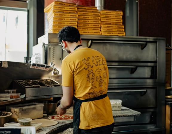 A chef from Scopa making pizzas with their back turned, wearing a yellow Scopa shirt.