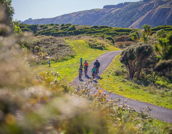 Four people ride bicycles on a path in Queen Elizabeth Regional Park in the Paraparaumu, New Zealand surrounded by nature.