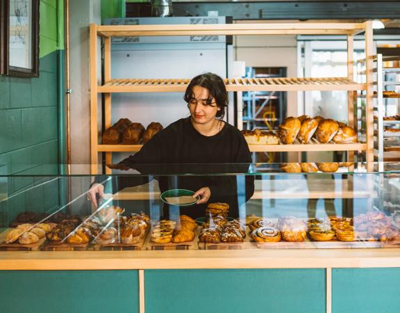 A baker uses tongs to pick up a pastry from the glass cabinet and put it on a plate.