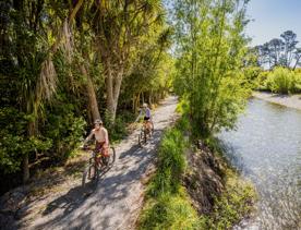 Two cyclists ride along the Waikanae River Trail on a sunny day.