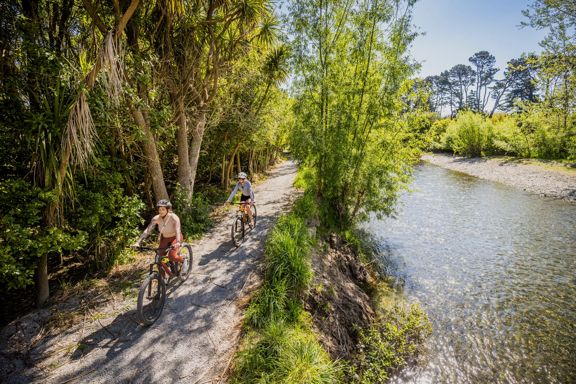 Two cyclists ride along the Waikanae River Trail on a sunny day.