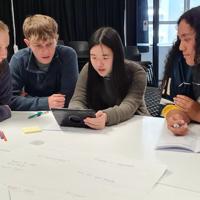 4 young adults sit at a table, one holds an iPad while the others look over their shoulder. Pends and paper are sprawled over the table.