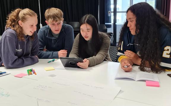 4 young adults sit at a table, one holds an iPad while the others look over their shoulder. Pends and paper are sprawled over the table.