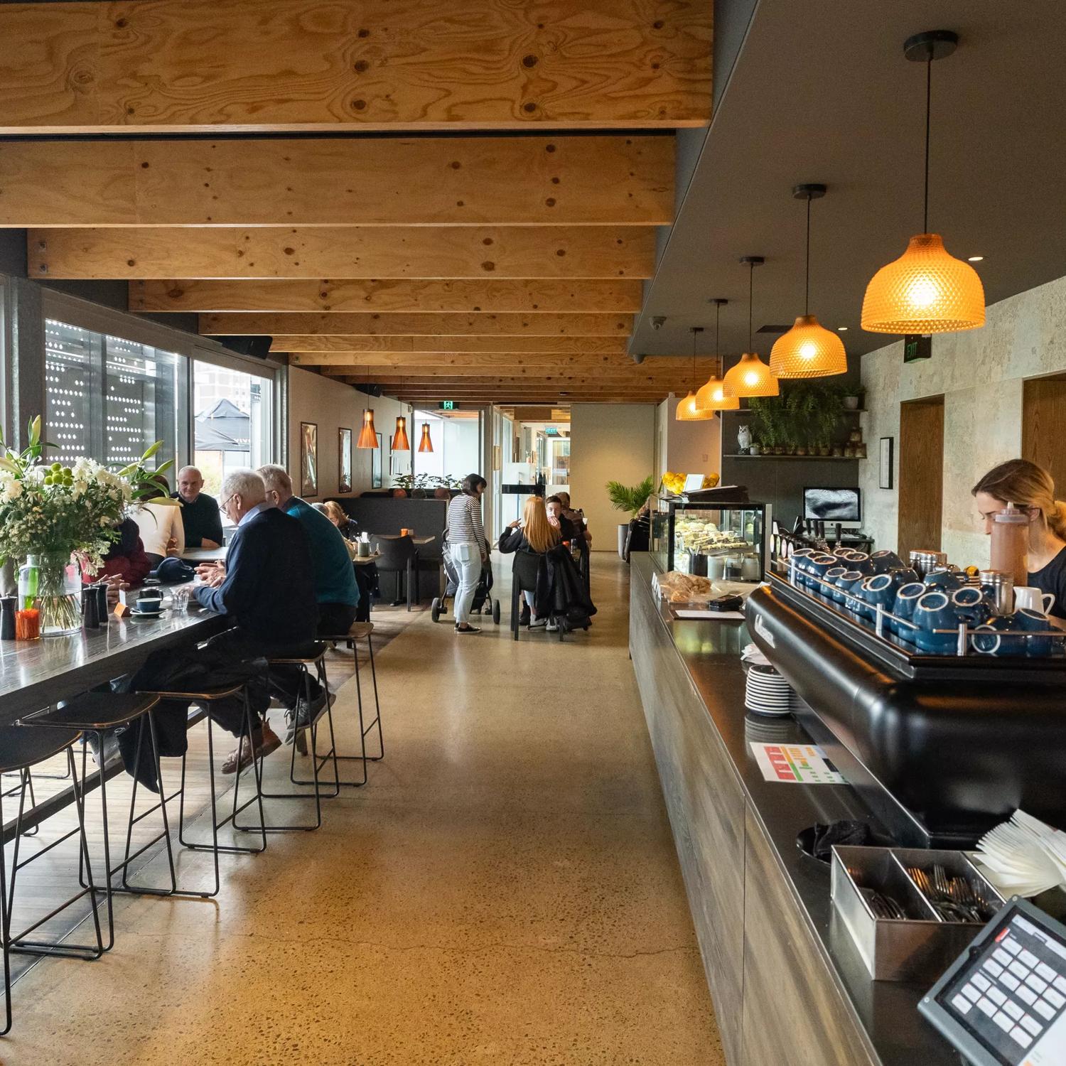 Wide look into the interior at Bellbird Eatery. A counter on the right with a barista at the coffee machine, and on the left, a long table with people drinking coffee.