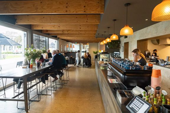 Wide look into the interior at Bellbird Eatery. A counter on the right with a barista at the coffee machine, and on the left, a long table with people drinking coffee.
