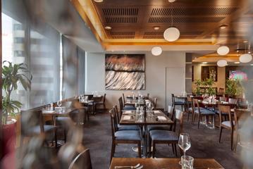 Inside a restaurant's dining room all set for service. There are wooden tables and chairs, grey carte and walls and small spherical lighting fixtures suspended from the ceiling.