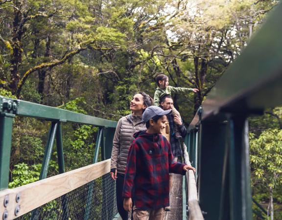 Two adults and two kids cross a bridge surrounded by trees on the Gentle Annie Track to Mount Holdsworth in Tararua Forest Park.