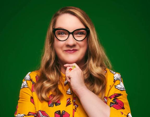 English comedian, writer and presenter, Sarah Millican, faces the camera with her hand to her chin and a subtle smile wearing a yellow floral blouse in front of a dark green wall. 