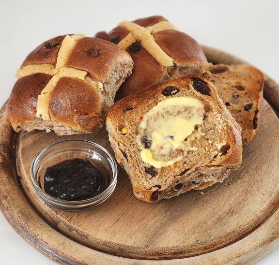 Three hot cross buns sitting on a round wooden bread board. One is cut open with a melting knob of butter sitting on it. To the left some jam sits in a glass ramekin. 