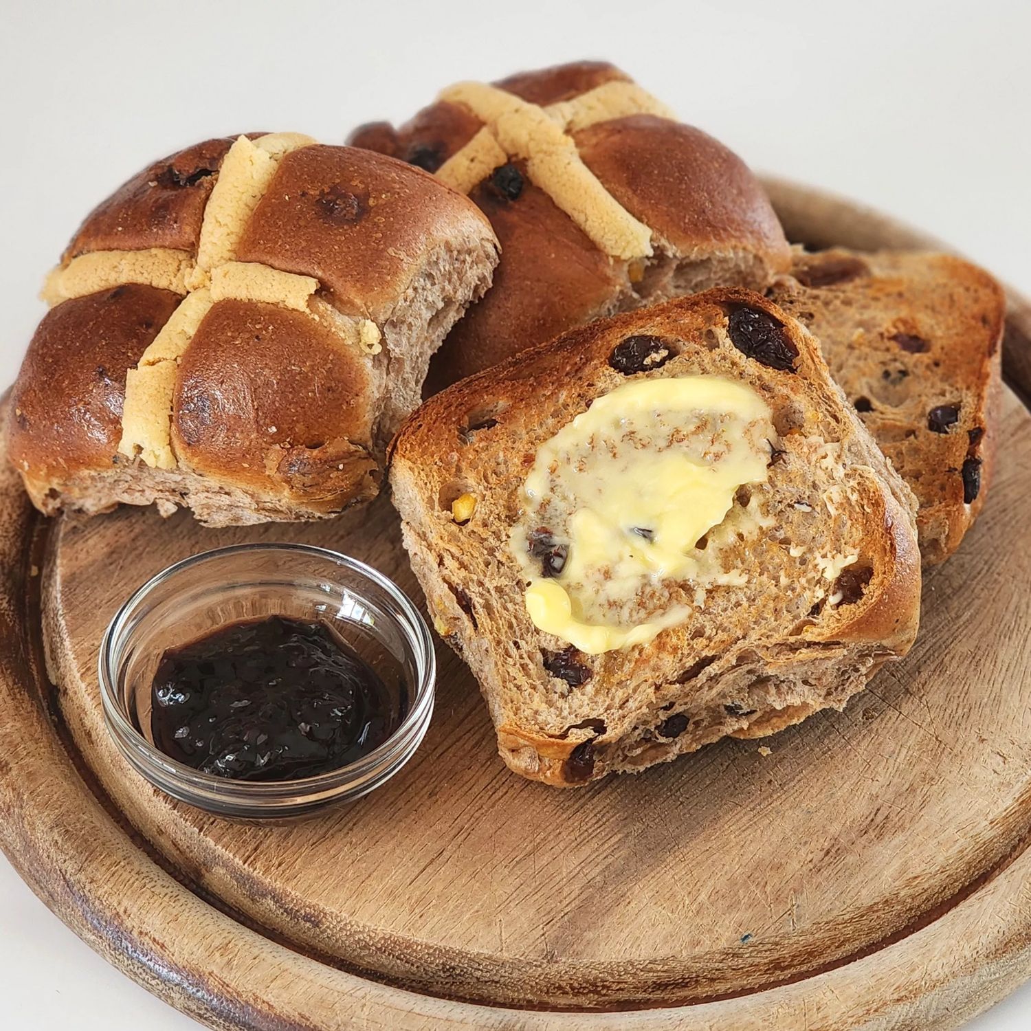 Three hot cross buns sitting on a round wooden bread board. One is cut open with a melting knob of butter sitting on it. To the left some jam sits in a glass ramekin.