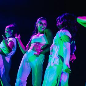 Five dancers wearing white under purple and turquoise lights.