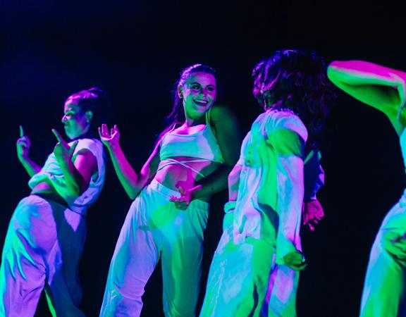 Five dancers wearing white under purple and turquoise lights.