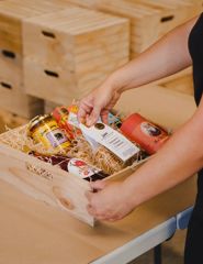 An open wooden box containing artisanal treats nestled into straw. An individual that mostly out of frame is placing a package of Balsamic Cheese Crackers into the box.