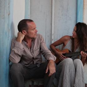 Damien Rice and a guitarist sit by a white and blue wall looking at one another.