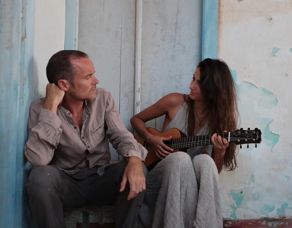 Damien Rice and a guitarist sit by a white and blue wall looking at one another.