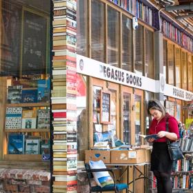 The exterior of Pegasus Books, with people outside reading, there is a large pillar made to look like thick books on the edge of the building.