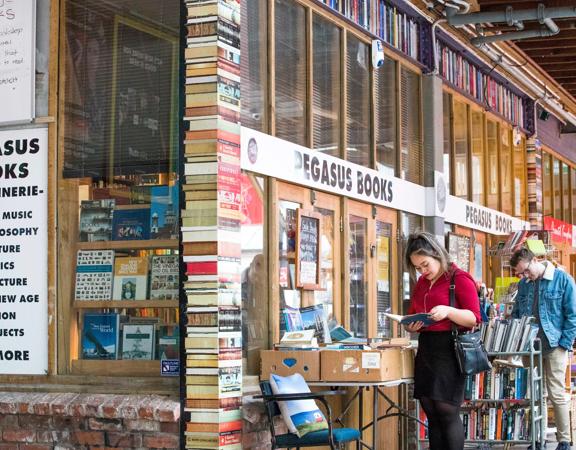 The exterior of Pegasus Books, with people outside reading, there is a large pillar made to look like thick books on the edge of the building.