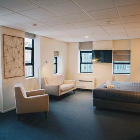 A room at the Park Hotel Lambton Quay has a bed with a grey cover against the right-hand wall, with two white chairs. Windows look out on the skyline.