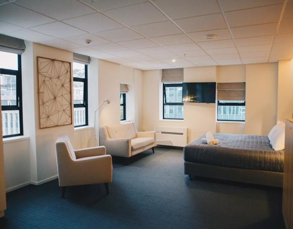 A room at the Park Hotel Lambton Quay has a bed with a grey cover against the right-hand wall, with two white chairs. Windows look out on the skyline.