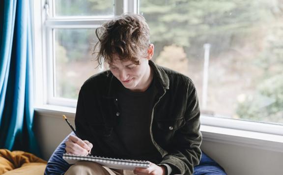 Wellington animator, Charlie Faulks, uses a pencil to draw in a sketchbook on his knee while sitting on a bean bag chair. 