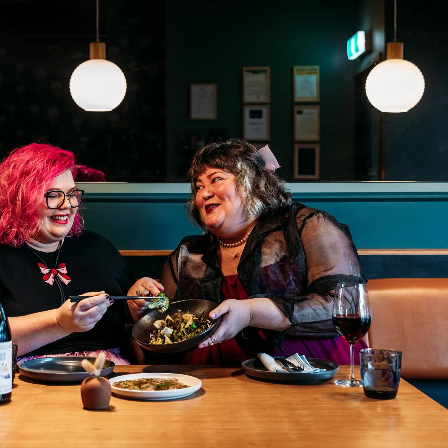 Two people sharing plates of food and a bottle of wine in Koji. the background is moody, with lantern lights and leather and blue velvet bench seats.