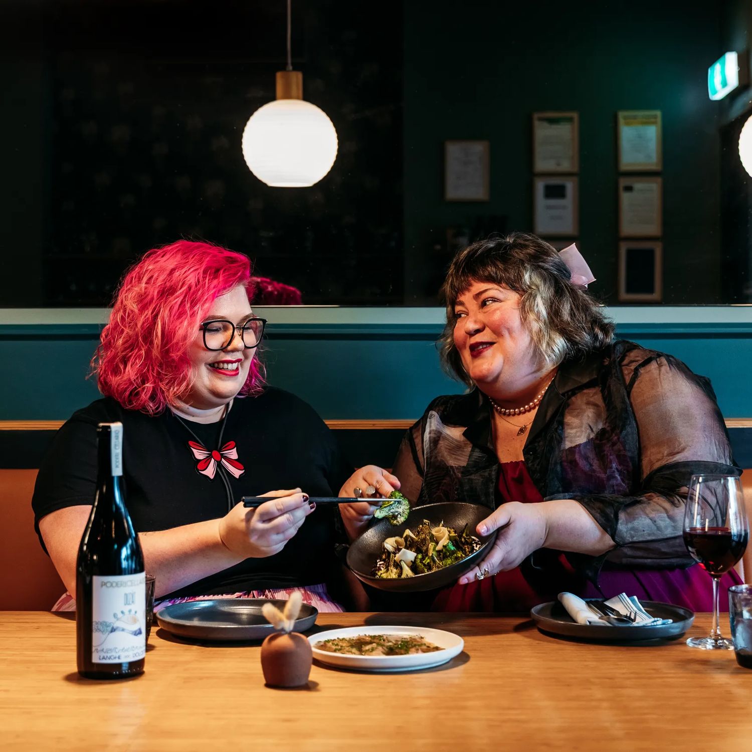 Two people sharing plates of food and a bottle of wine in Koji. the background is moody, with lantern lights and leather and blue velvet bench seats.