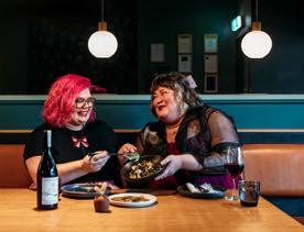 Two people sharing plates of food and a bottle of wine in Koji. the background is moody, with lantern lights and leather and blue velvet bench seats.