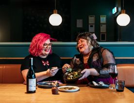 Two people sharing plates of food and a bottle of wine in Koji. the background is moody, with lantern lights and leather and blue velvet bench seats.