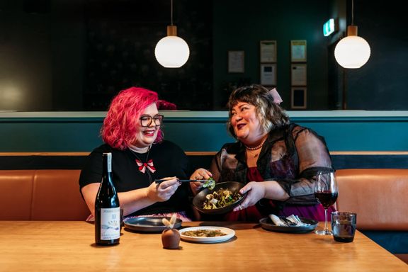 Two people sharing plates of food and a bottle of wine in Koji. the background is moody, with lantern lights and leather and blue velvet bench seats.