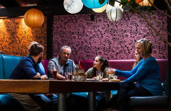 A family of six sit a a booth, having food and drinks at a restaurant with floral wallpaper and paper lanterns. 