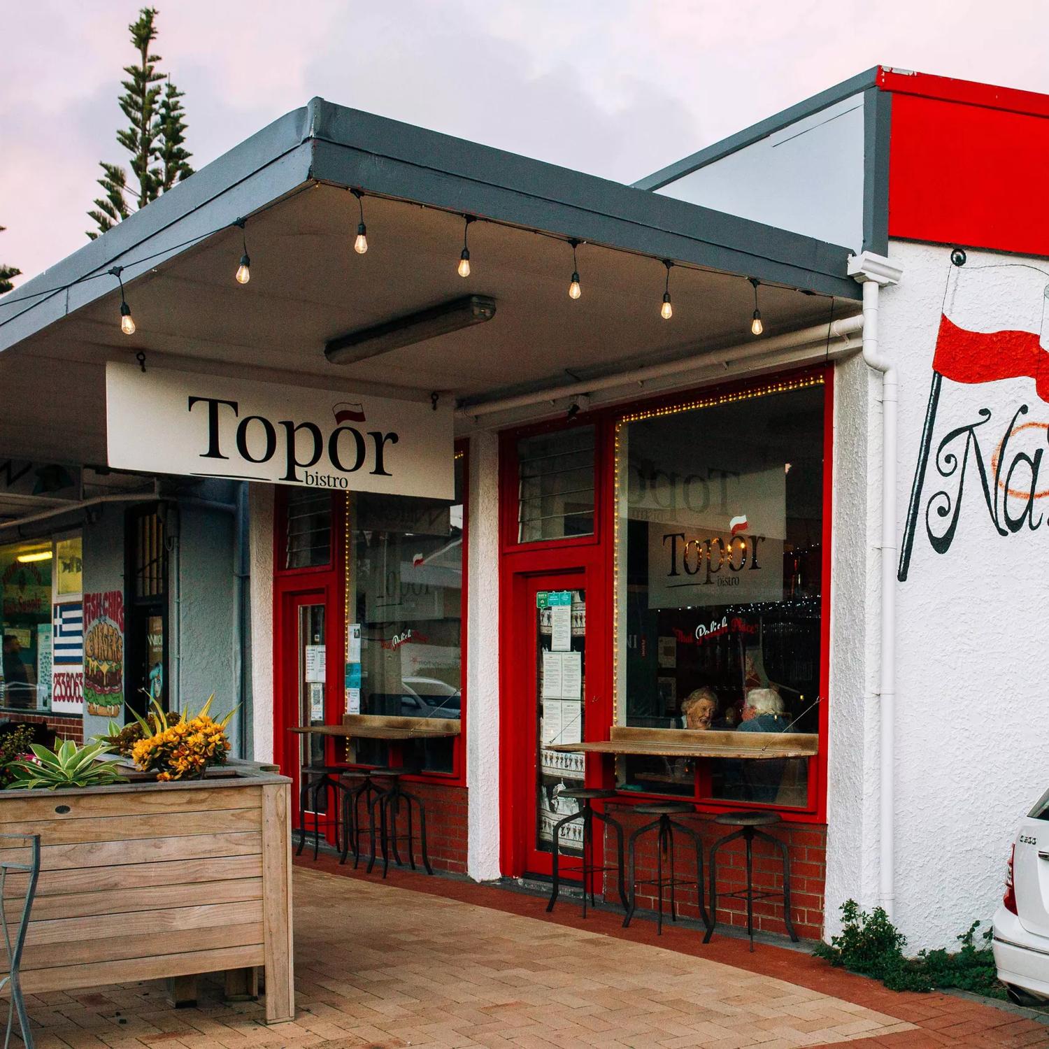 The exterior of Topor Bistro, a Polish restaurant in Porirua, New Zealand. The small white building has hanging lights and red accents. 