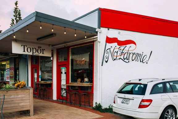 The exterior of Topor Bistro, a Polish restaurant in Porirua, New Zealand. The small white building has hanging lights and red accents.
