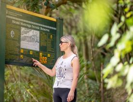 A person reading the Hemi Matenga Scenic reserve sign, with bush in the background.