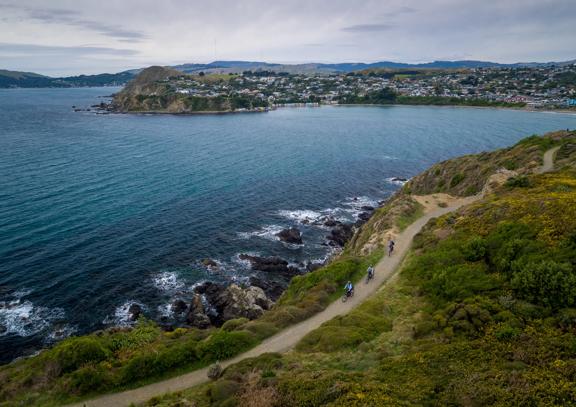 A birdseye view of three cyclists riding along Te Onepoto Loop Track in Whitireia Park along the coast in Porirua, New Zealand. 