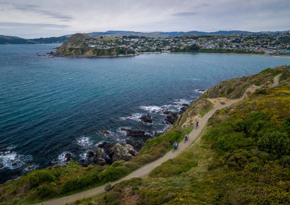 A birdseye view of three cyclists riding along Te Onepoto Loop Track in Whitireia Park along the coast in Porirua, New Zealand.