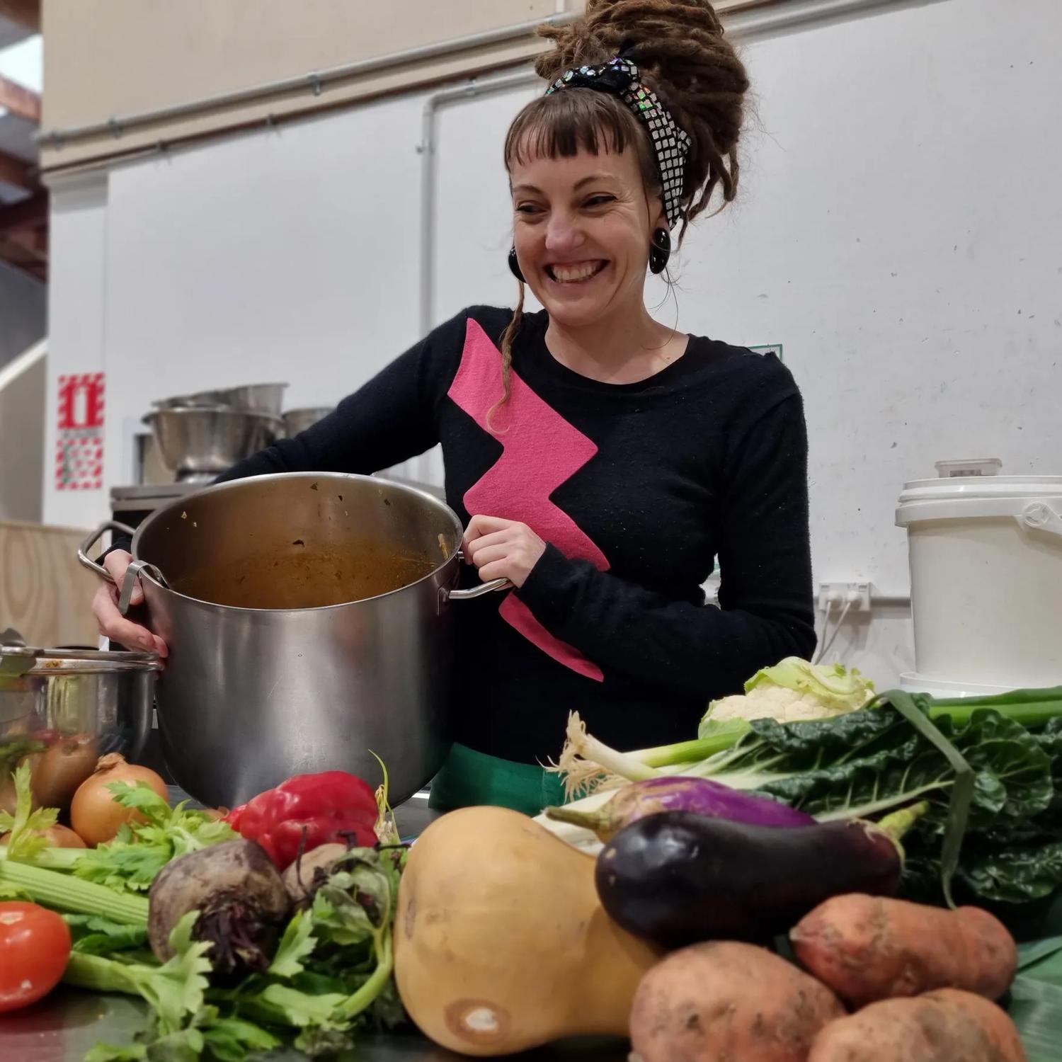A smiling person holds a large cooking pot while standing by a table covered in raw vegetables. 