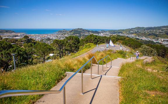 Looking down a stretch of concrete steps with a silver handrail on top of Mount Victoria, on a sunny blue-sky day. Wellington's southern suburbs are in the distance along with the airport on the left, and the Cook Strait.