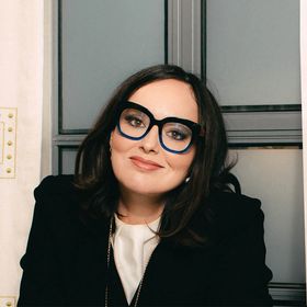 Deborah Frances-White with a subtle smile in front of an industrial window.