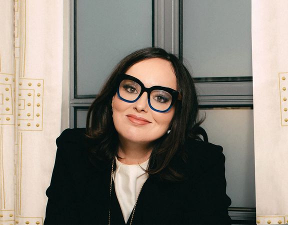 Deborah Frances-White with a subtle smile in front of an industrial window.
