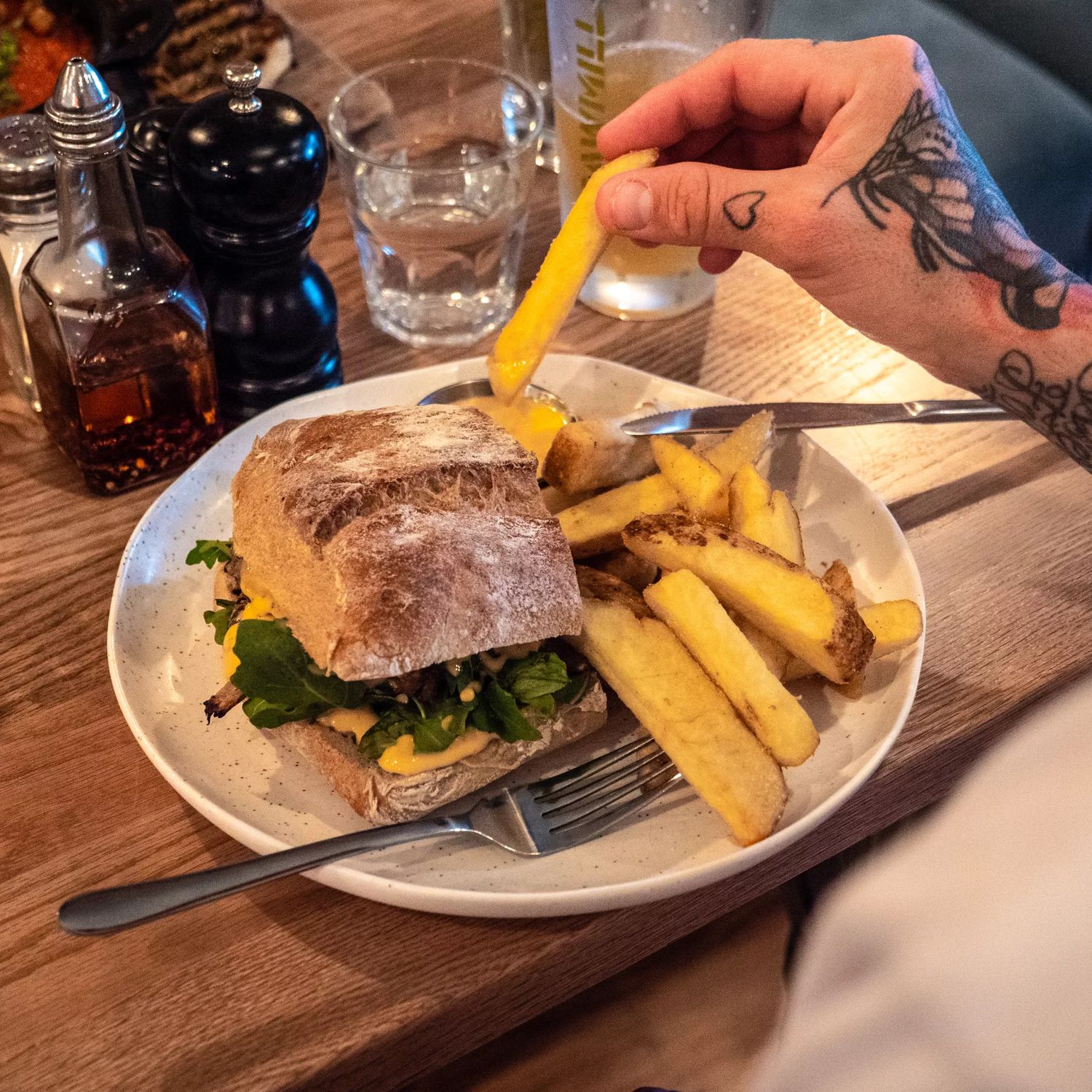 Sandwich with thick-cut fries on a white plate, with a tattooed hand reaching for a fry at a restaurant table.