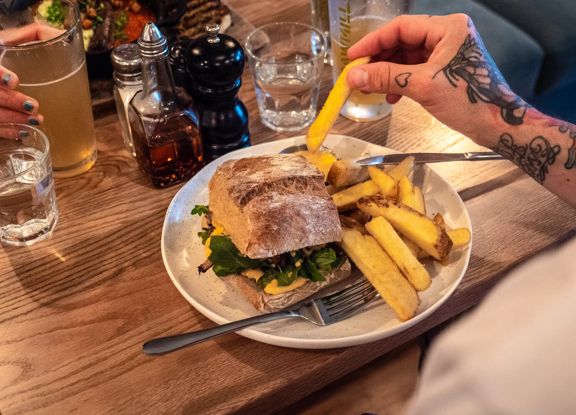 Sandwich with thick-cut fries on a white plate, with a tattooed hand reaching for a fry at a restaurant table.