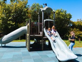 Two kids use a double slide at a playground with an adult supervising.