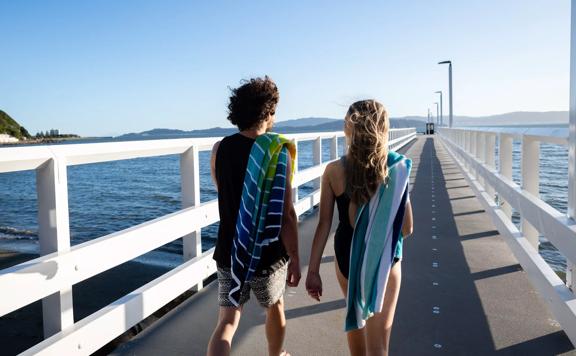 2 people walk along the pier at Days Bay in their swimsuits with towels over their shoulders.