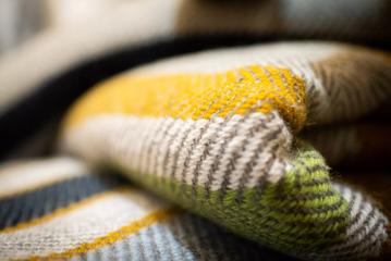 A close-up of a folded knit blanket. The blanket is light grey with yellow, green and navy blue stripes.