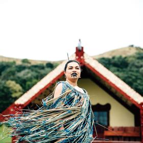 A Māori person standing in front of a wharenui on a windy day.