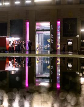The City Gallery Wellington's front entrance at night with an illuminated small red caravan parked on the left and five people queued in front of it. The lights from the gallery and caravan are reflected in the water's surface in the foreground. 